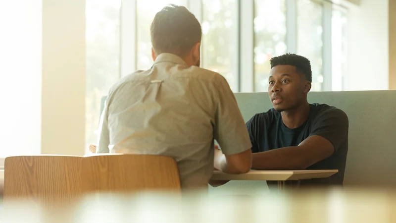 Two colleagues in a coffee shop having an honest conversation, one confiding something personal while the other listens attentively