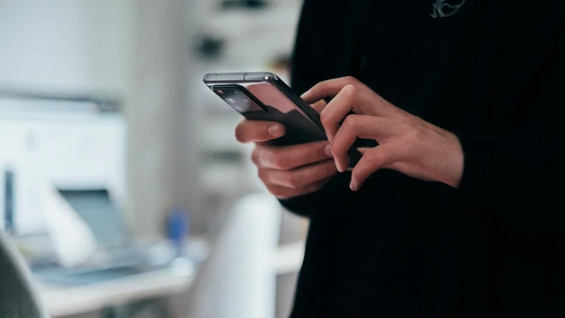 Person sitting in bed at night, face lit by phone screen, empty glass on bedside table in soft focus