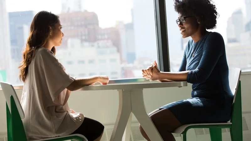 Dos personas en una conversación sincera y directa — una inclinándose hacia la otra, ambas visiblemente aliviadas; la experiencia del impostor pierde su poder a través de la conexión y el reconocimiento compartido