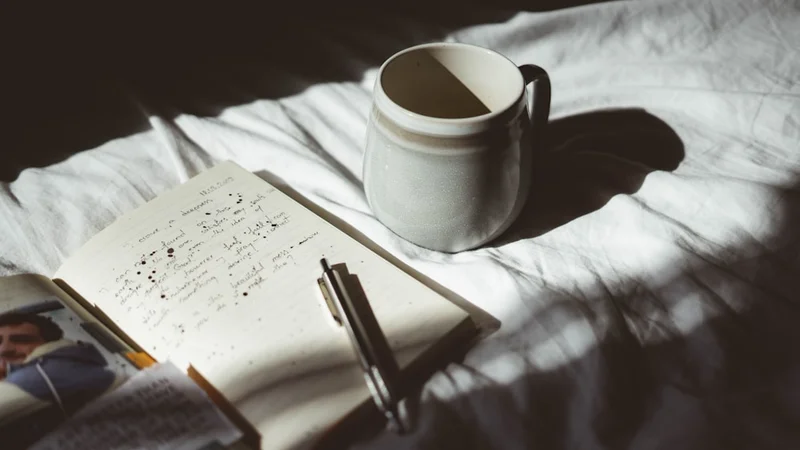 Mains enveloppant une tasse blanche, un journal ouvert sur une table en bois avec la lumière du matin entrant par la fenêtre