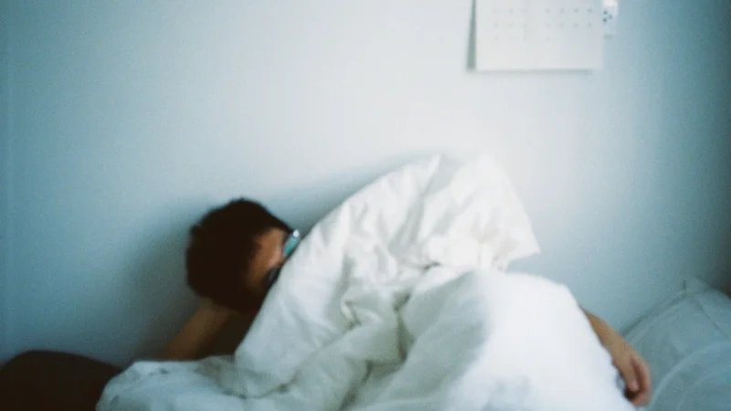 Persona seduta sul bordo del letto alla luce del mattino, che si sfrega gli occhi, con una sveglia che mostra le 8:00 sullo sfondo — espressione di stanchezza nonostante una notte intera di sonno