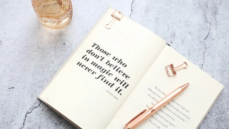 pessoa escrevendo em um caderno de capa dura em uma mesa de madeira com luz da manhã entrando pela janela e um café preto ao lado